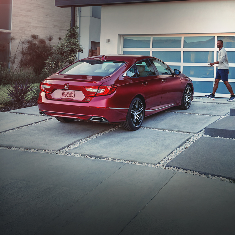 Seven-eighth rear passenger-side view of the 2021 Accord Touring 2.0T, shown in Radiant Red Metallic, parked on a driveway in front of a modern family home. 